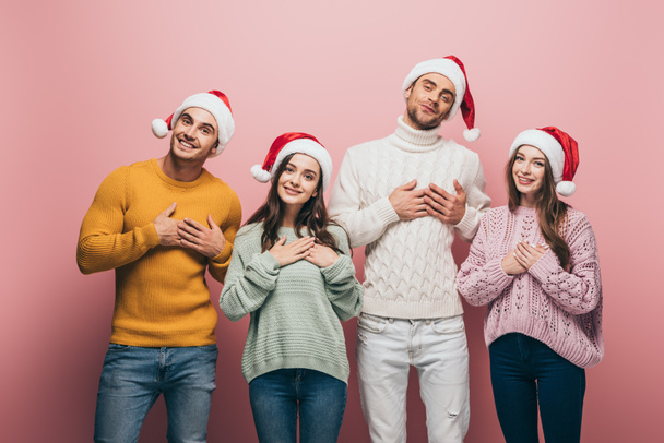 happy friends in sweaters and santa hats holding hands on hearts, isolated on pink - Fotoğraf, Görsel