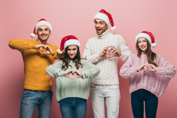 happy friends in sweaters and santa hats showing heart symbols, isolated on pink - Photo, Image