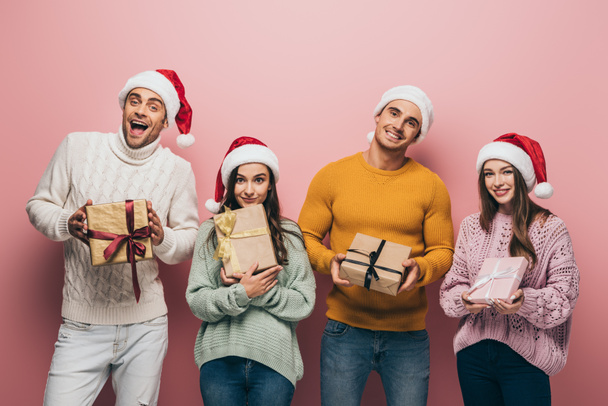 cheerful friends in santa hats holding christmas presents, isolated on pink - Zdjęcie, obraz