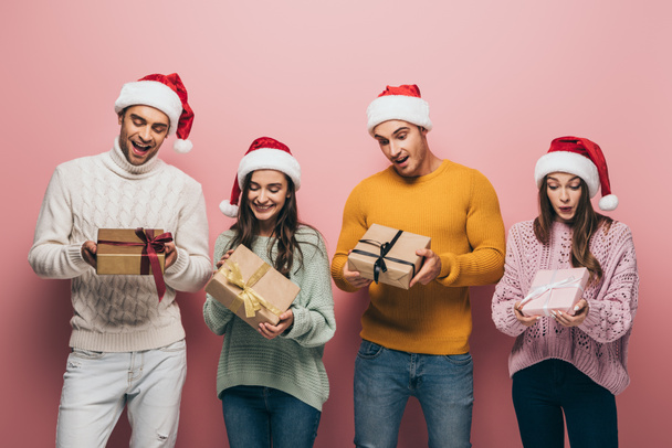beautiful surprised friends in santa hats holding christmas presents, isolated on pink - Fotoğraf, Görsel