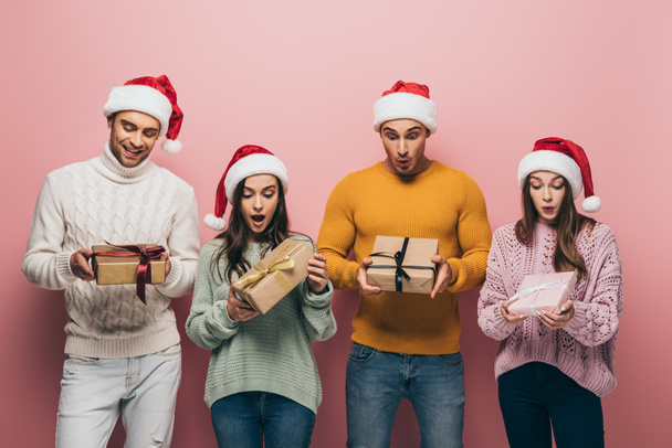 surprised friends in santa hats holding christmas presents, isolated on pink - Photo, Image