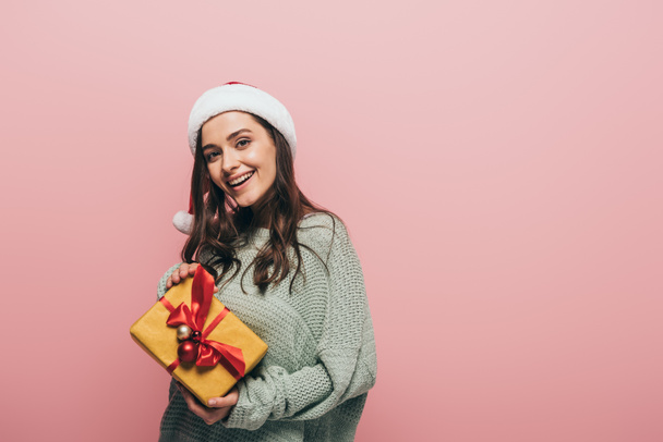 smiling girl in sweater and santa hat holding christmas gift, isolated on pink - Valokuva, kuva