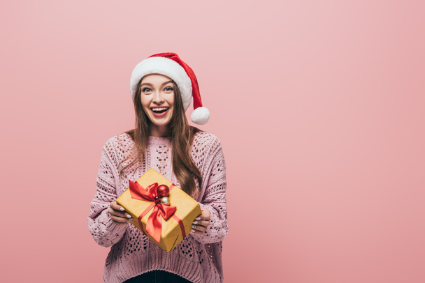 smiling woman in sweater and santa hat holding christmas gift, isolated on pink - Fotó, kép