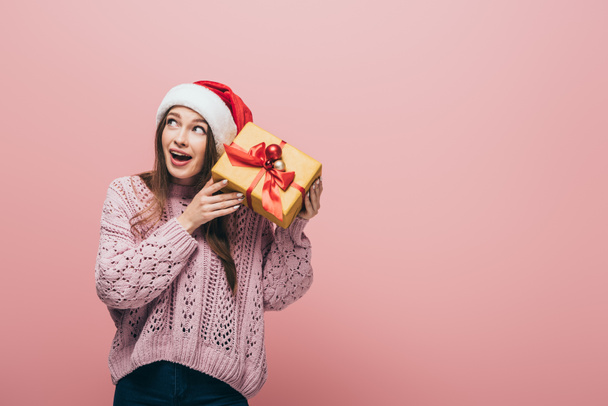 emotional woman in sweater and santa hat holding christmas gift, isolated on pink - Fotografie, Obrázek