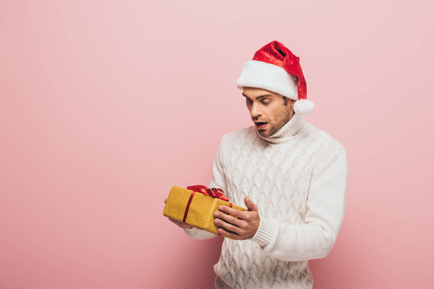 σοκαρισμένος άντρας με πουλόβερ και καπέλο Σάντα κρατώντας χριστουγεννιάτικο δώρο, απομονωμένος σε ροζ - Φωτογραφία, εικόνα
