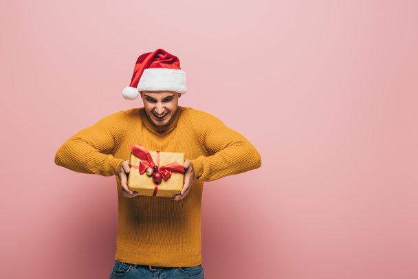 smiling man in santa hat holding christmas gift box, isolated on pink - Foto, imagen