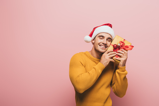 smiling man in sweater and santa hat holding christmas gift box, isolated on pink - Foto, imagen