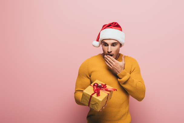 shocked man in sweater and santa hat looking at christmas present, isolated on pink - Φωτογραφία, εικόνα