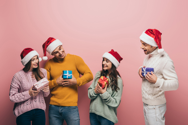 happy friends in santa hats holding christmas presents, isolated on pink - Photo, Image
