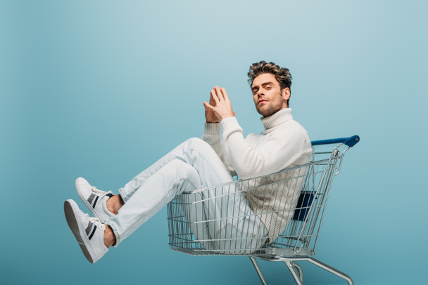 handsome pensive man sitting in shopping cart, isolated on blue - Foto, immagini