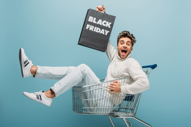 excited man sitting in shopping cart while holding shopping bag with black friday sign, isolated on blue - Foto, Bild