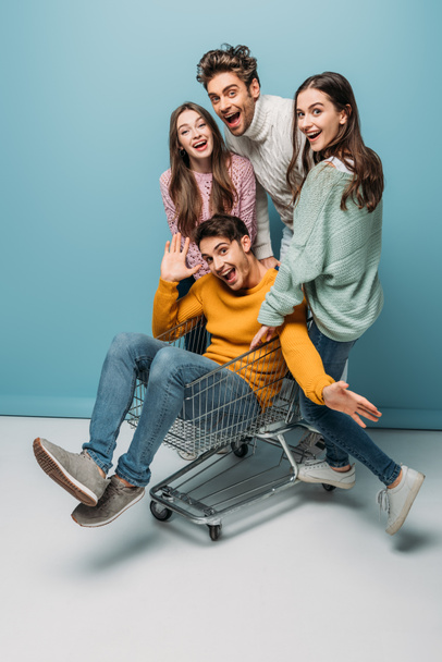 happy friends having fun and riding in shopping cart on blue - Foto, afbeelding