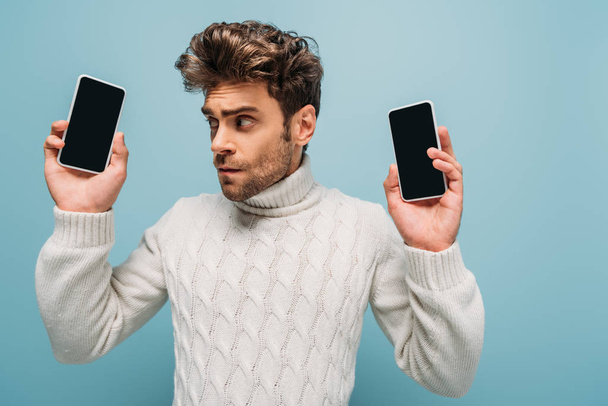 emotional man looking at two smartphones with blank screens, isolated on blue - Foto, Imagem