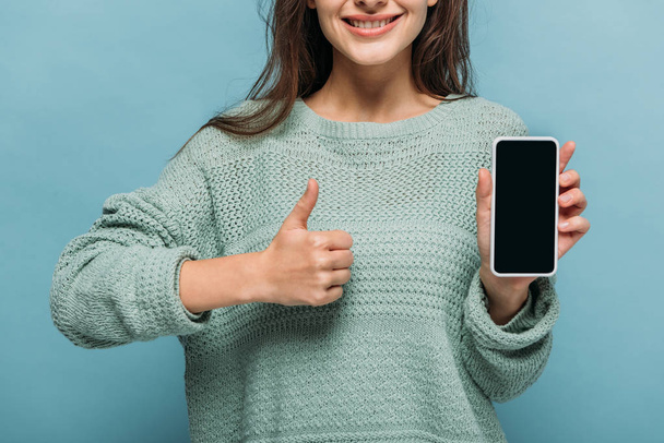 cropped view of smiling woman showing thumb up and smartphone with blank screen, isolated on blue - Photo, Image