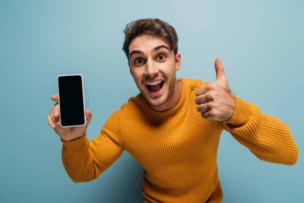 excited young man showing thumb up and smartphone with blank screen, isolated on blue - Foto, Imagem
