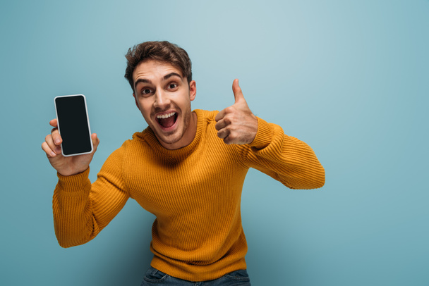 excited man showing thumb up and smartphone with blank screen, isolated on blue - Фото, изображение