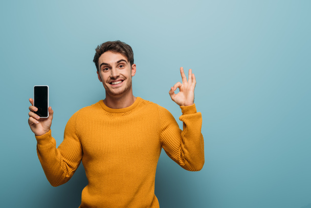 positive man showing ok sign and smartphone with blank screen, isolated on blue - Foto, Bild