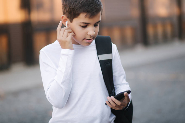 Teenage boy take out a wireless headphones and put then into the ears. Young boy in white shirt with backpack. Boy listen music - Fotoğraf, Görsel