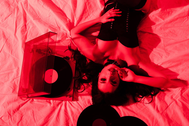 attractive passionate woman lying on bed with vinyl records in red light - Foto, imagen