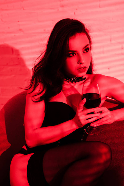 sensual woman sitting in armchair with glass of wine in red light  - Fotoğraf, Görsel