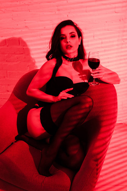 beautiful passionate woman sitting in armchair with glass of wine in red light  - Photo, Image