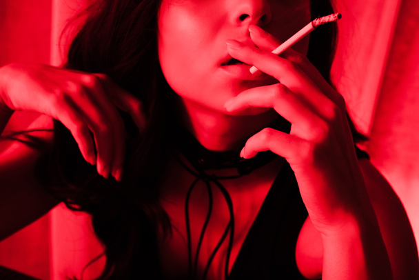 cropped view of attractive sexy girl smoking cigarette in red light - Fotografie, Obrázek