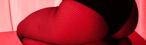 recortado vista de sexy nalgas de chica en medias de rejilla en la luz roja, tiro panorámico
 - Foto, Imagen
