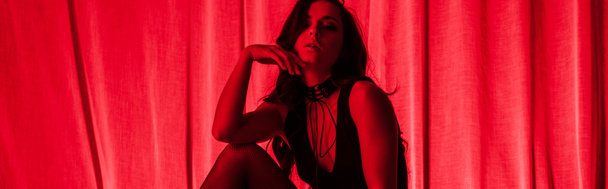 panoramic shot of sensual young woman posing in bodysuit and fishnet tights in red light - Photo, Image