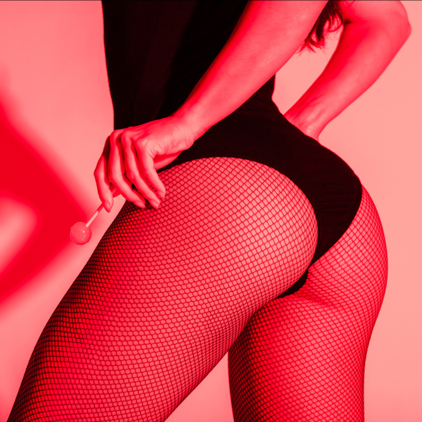 cropped view of sexy buttocks of girl in fishnet tights holding lollipop in red light - Photo, Image