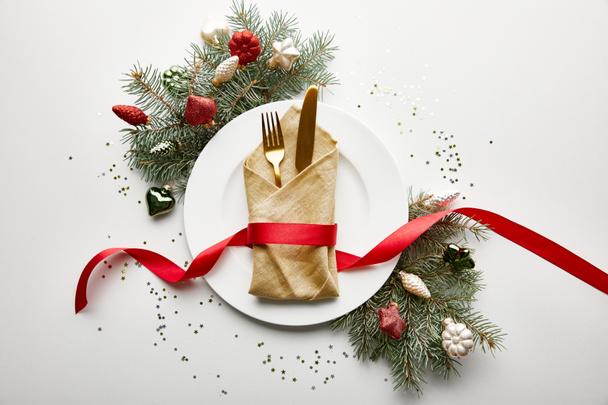 top view of white plate with napkin, cutlery and ribbon near festive Christmas tree branch with baubles and confetti on white background - Photo, Image