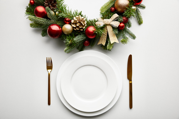 vue de dessus des assiettes blanches avec des couverts dorés près de couronne de Noël festive avec des boules sur fond blanc
 - Photo, image
