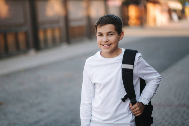Портрет счастливого улыбающегося подростка в белой толстовке с рюкзаком на улице
 - Фото, изображение