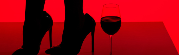 vista cortada de silhueta preta de menina sensual em saltos com copo de vinho isolado em vermelho, tiro panorâmico
 - Foto, Imagem