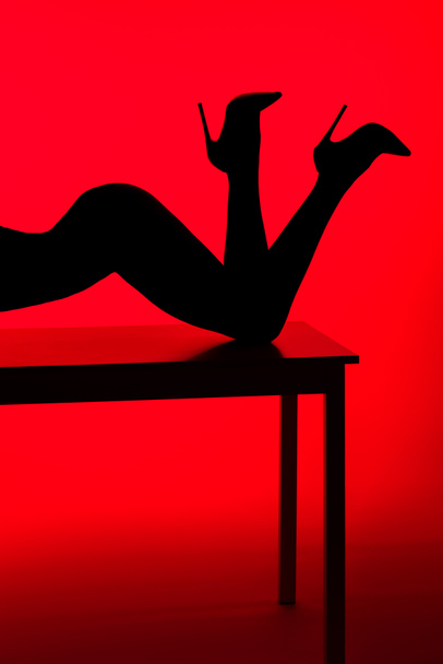 black silhouette of passionate girl in heels posing on table isolated on red - Zdjęcie, obraz