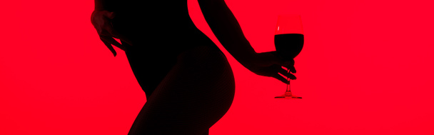 cropped view of black silhouette of sexy girl holding glass of wine isolated on red, panoramic shot - Fotó, kép
