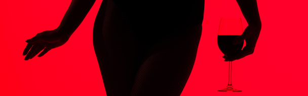 cropped view of black silhouette of sexy girl holding glass of wine isolated on red, panoramic shot - Foto, afbeelding