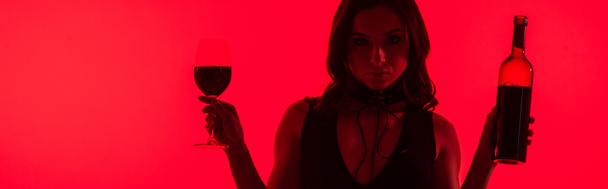 panoramic shot of attractive sexy woman holding bottle and glass of wine isolated on red - Photo, Image
