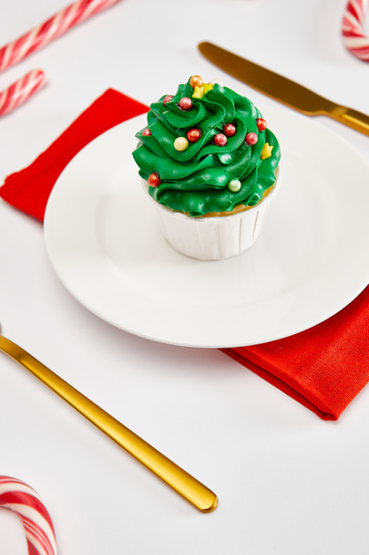delicioso cupcake en plato blanco con cubiertos de oro, caramelos y servilleta roja en la superficie blanca
 - Foto, Imagen