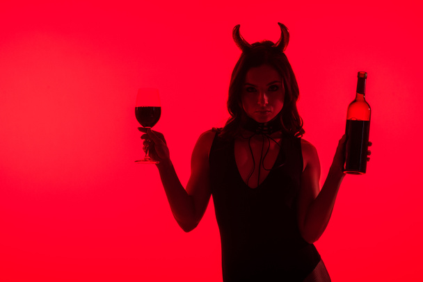 sensual girl in Devil costume holding glass and bottle of wine, isolated on red - Foto, afbeelding