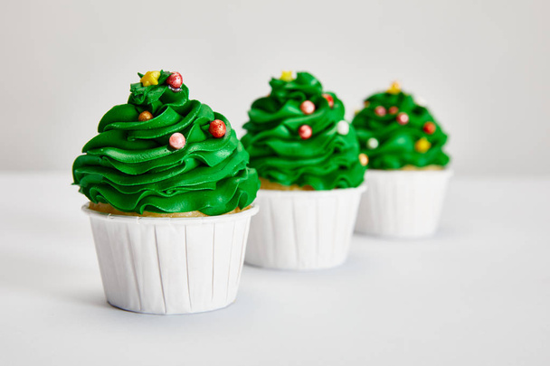 selective focus of tasty Christmas tree cupcakes in row on white surface isolated on grey - Foto, Imagen