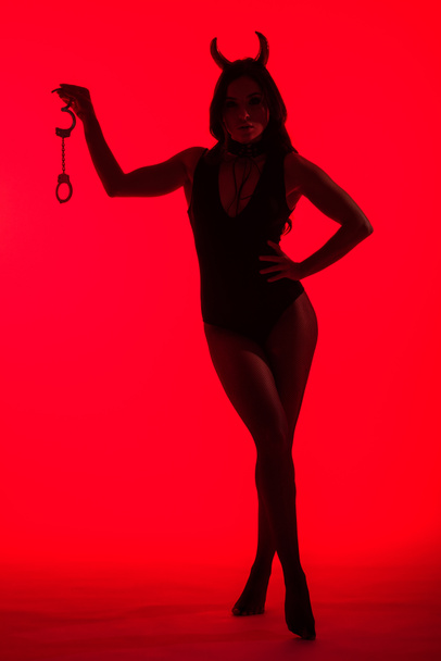 silhouette of sexy woman in Devil costume holding handcuffs, isolated on red - Фото, изображение