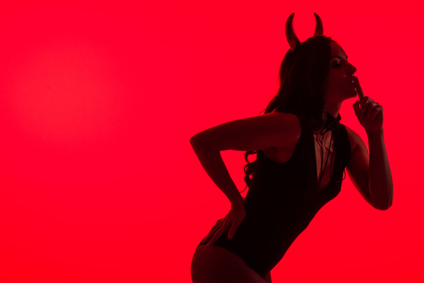 Silhouette einer sexy Frau im Teufelskostüm, die das Symbol der Stille zeigt, isoliert auf Rot - Foto, Bild