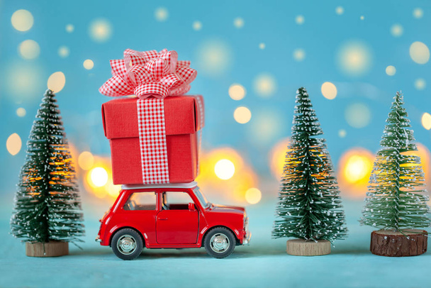 Red car carrying on roof a gift box and Christmas tree on a blue background. New Year and Christmas concept. - Fotó, kép