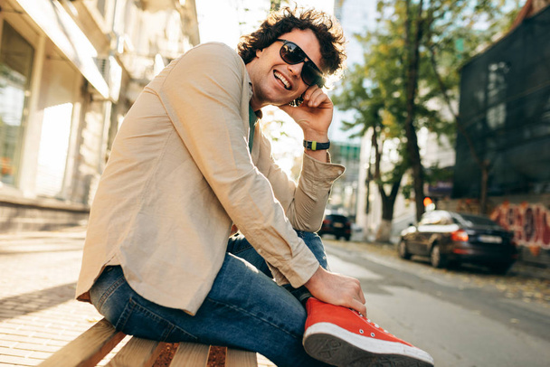 カールした髪をした幸せな笑顔の男は、街の通りの屋外のベンチに座っている社会的な広告のポーズ。興奮陽気な学生の男は外で休む. - 写真・画像