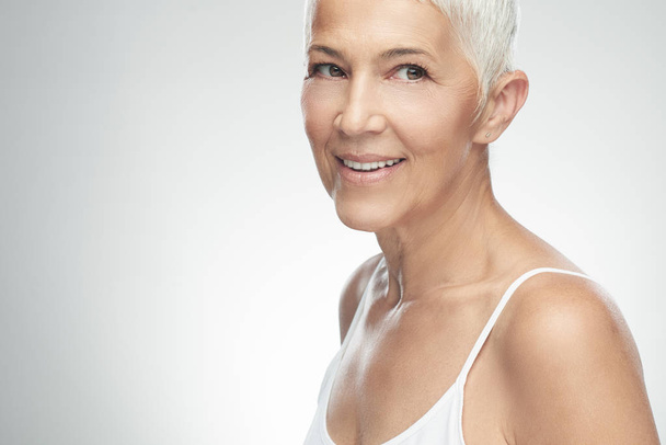 Beautiful smiling senior woman with short gray hair posing in front of gray background. Beauty photography. - Fotografie, Obrázek