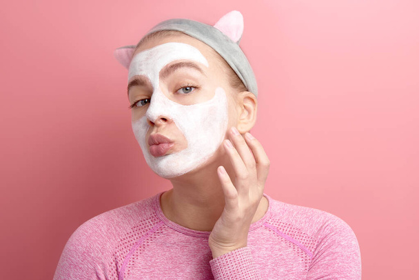 young woman applying white clay cosmetic mask, touches face looking at camera as in mirror, home facial care, female morning and evening routine - Photo, Image