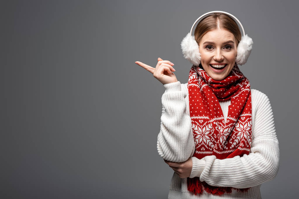 attractive excited girl in white sweater, earmuffs and scarf pointing at something, isolated on grey - Foto, Imagen