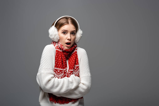 attractive shocked girl in white sweater, earmuffs and scarf posing with crossed arms, isolated on grey - Photo, Image