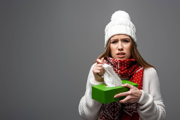 upset sick girl in knitted hat holding paper napkins, isolated on grey - Foto, imagen