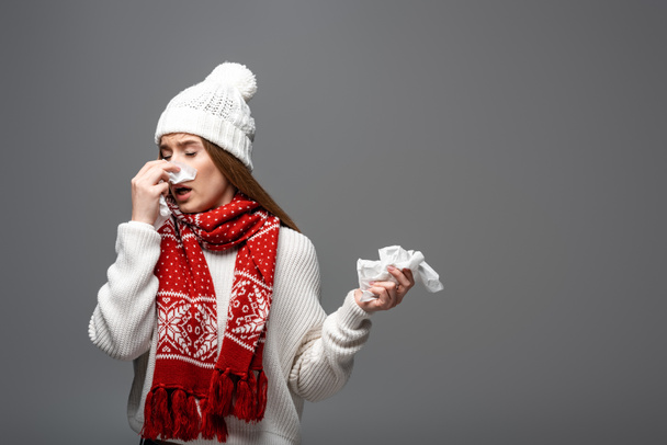 sick woman in knitted hat and scarf having runny nose with paper napkins, isolated on grey - Photo, Image